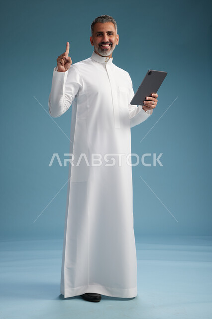 بورتريه لرجل عربي سعودي  خليجي يرتدي الثوب  الابيض يستخدم جهازاً لوحياً رقمياً, كامل الجسم, خلفيه زرقاء