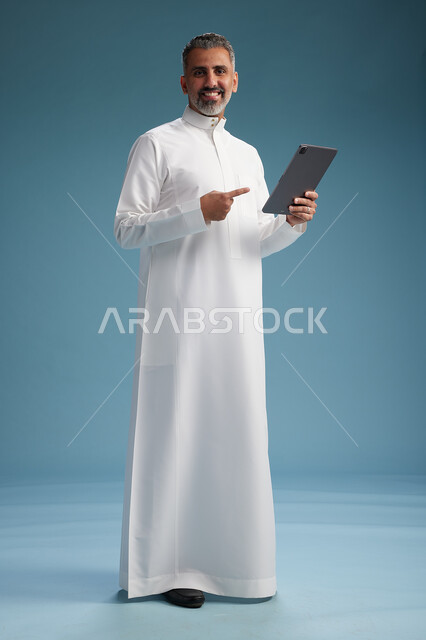 A portrait of a Saudi Arabian Gulf man wearing a white thobe using a digital tablet, full body, with a blue background.