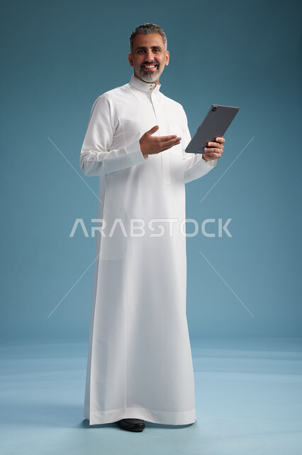 بورتريه لرجل عربي سعودي  خليجي يرتدي الثوب  الابيض يستخدم جهازاً لوحياً رقمياً, كامل الجسم, خلفيه زرقاء