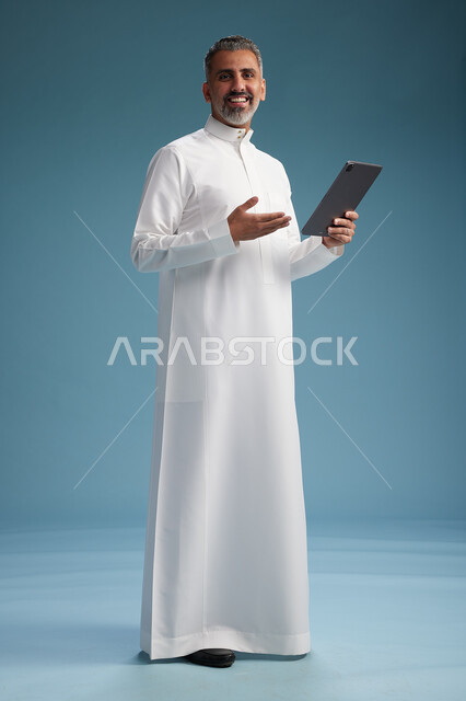 A portrait of a Saudi Arabian Gulf man wearing a white thobe using a digital tablet, full body, with a blue background.