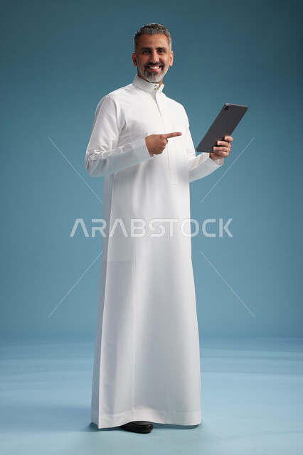 بورتريه لرجل عربي سعودي  خليجي يرتدي الثوب  الابيض يستخدم جهازاً لوحياً رقمياً, كامل الجسم, خلفيه زرقاء