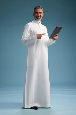 A portrait of a Saudi Arabian Gulf man wearing a white thobe using a digital tablet, full body, with a blue background.
