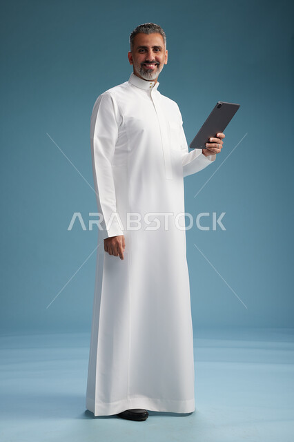 بورتريه لرجل عربي سعودي  خليجي يرتدي الثوب  الابيض يستخدم جهازاً لوحياً رقمياً, كامل الجسم, خلفيه زرقاء