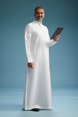 A portrait of a Saudi Arabian Gulf man wearing a white thobe using a digital tablet, full body, with a blue background.