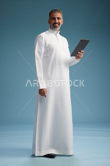 بورتريه لرجل عربي سعودي  خليجي يرتدي الثوب  الابيض يستخدم جهازاً لوحياً رقمياً, كامل الجسم, خلفيه زرقاء