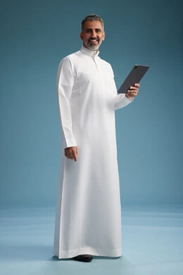 A portrait of a Saudi Arabian Gulf man wearing a white thobe using a digital tablet, full body, with a blue background. A portrait of a Saudi Arabian Gulf man wearing a white thobe using a digital tablet, full body, with a blue background.