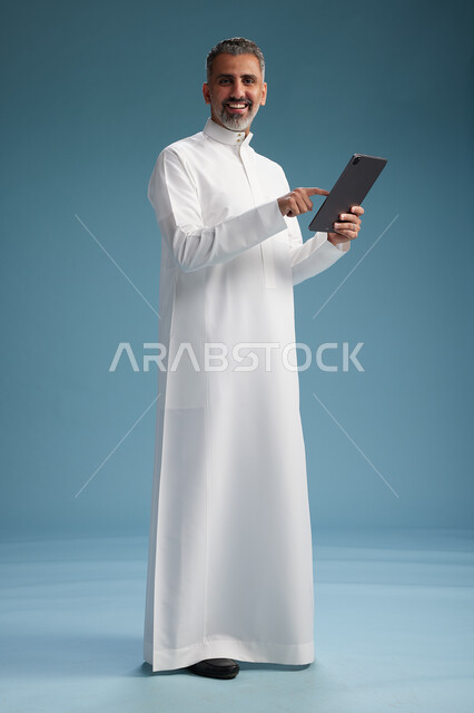 بورتريه لرجل عربي سعودي  خليجي يرتدي الثوب  الابيض يستخدم جهازاً لوحياً رقمياً, كامل الجسم, خلفيه زرقاء