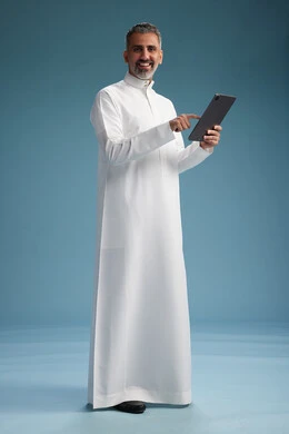 بورتريه لرجل عربي سعودي  خليجي يرتدي الثوب  الابيض يستخدم جهازاً لوحياً رقمياً, كامل الجسم, خلفيه زرقاء
