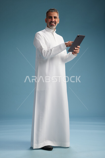 بورتريه لرجل عربي سعودي  خليجي يرتدي الثوب  الابيض يستخدم جهازاً لوحياً رقمياً, كامل الجسم, خلفيه زرقاء