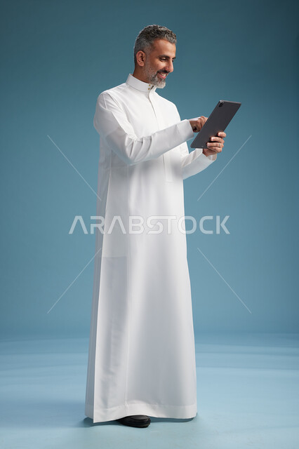 بورتريه لرجل عربي سعودي  خليجي يرتدي الثوب  الابيض يستخدم جهازاً لوحياً رقمياً, كامل الجسم, خلفيه زرقاء