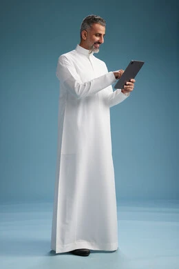 A portrait of a Saudi Arabian Gulf man wearing a white thobe using a digital tablet, full body, with a blue background.
