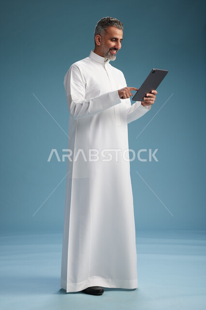 بورتريه لرجل عربي سعودي  خليجي يرتدي الثوب  الابيض يستخدم جهازاً لوحياً رقمياً, كامل الجسم, خلفيه زرقاء