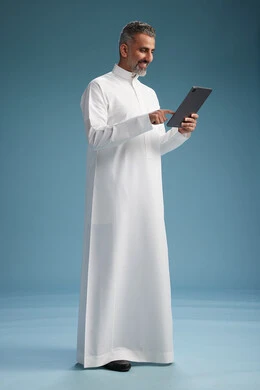 بورتريه لرجل عربي سعودي  خليجي يرتدي الثوب  الابيض يستخدم جهازاً لوحياً رقمياً, كامل الجسم, خلفيه زرقاء