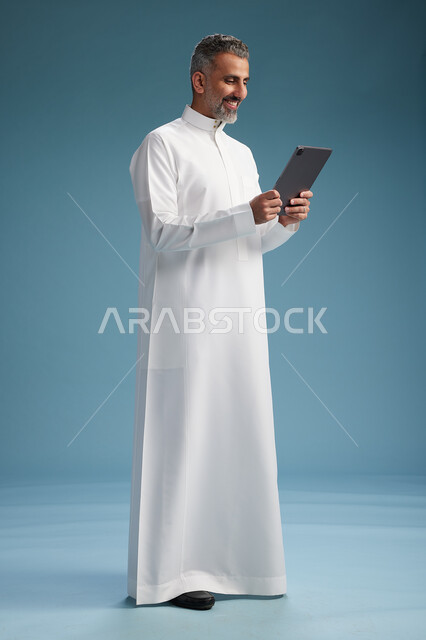 بورتريه لرجل عربي سعودي  خليجي يرتدي الثوب  الابيض يستخدم جهازاً لوحياً رقمياً, كامل الجسم, خلفيه زرقاء