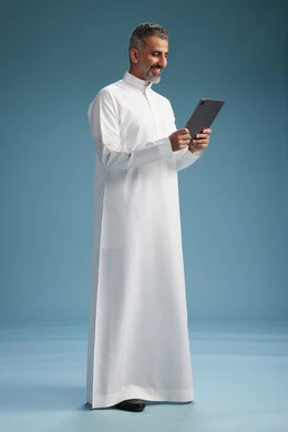 بورتريه لرجل عربي سعودي  خليجي يرتدي الثوب  الابيض يستخدم جهازاً لوحياً رقمياً, كامل الجسم, خلفيه زرقاء