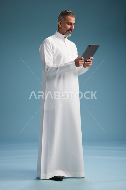 بورتريه لرجل عربي سعودي  خليجي يرتدي الثوب  الابيض يستخدم جهازاً لوحياً رقمياً, كامل الجسم, خلفيه زرقاء