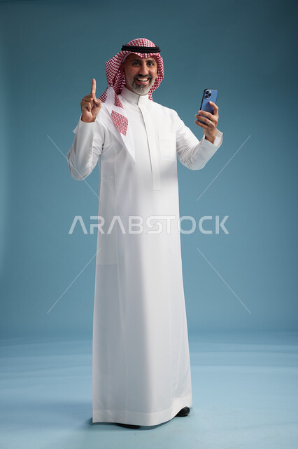 رجل  عربي سعودي خليجي يرتدي الثوب الابيض والشماغ  يستخدم هاتفاً ذكياً, الجسم بالطول الكامل, خلفيه زرقاء
