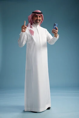 A Saudi Arabian Gulf Arab man wearing a white thobe and a shemagh is using a smartphone, full-body, with a blue background. A Saudi Arabian Gulf Arab man wearing a white thobe and a shemagh is using a smartphone, full-body, with a blue background.