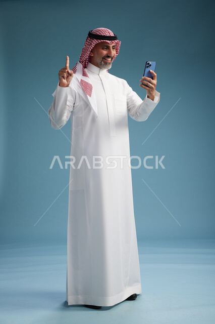 رجل  عربي سعودي خليجي يرتدي الثوب الابيض والشماغ  يستخدم هاتفاً ذكياً, الجسم بالطول الكامل, خلفيه زرقاء