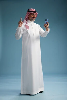 A Saudi Arabian Gulf Arab man wearing a white thobe and a shemagh is using a smartphone, full-body, with a blue background. A Saudi Arabian Gulf Arab man wearing a white thobe and a shemagh is using a smartphone, full-body, with a blue background.