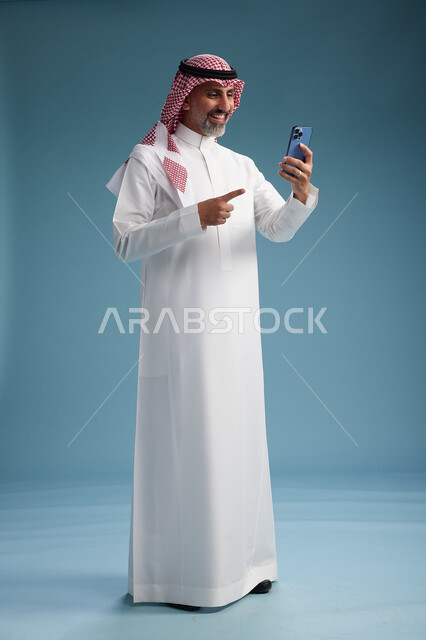 رجل  عربي سعودي خليجي يرتدي الثوب الابيض والشماغ  يستخدم هاتفاً ذكياً, الجسم بالطول الكامل, خلفيه زرقاء