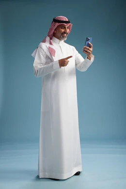 A Saudi Arabian Gulf Arab man wearing a white thobe and a shemagh is using a smartphone, full-body, with a blue background.