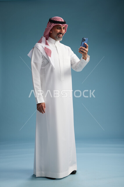 رجل  عربي سعودي خليجي يرتدي الثوب الابيض والشماغ  يستخدم هاتفاً ذكياً, الجسم بالطول الكامل, خلفيه زرقاء