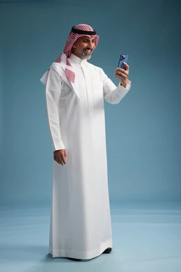 A Saudi Arabian Gulf Arab man wearing a white thobe and a shemagh is using a smartphone, full-body, with a blue background. A Saudi Arabian Gulf Arab man wearing a white thobe and a shemagh is using a smartphone, full-body, with a blue background.