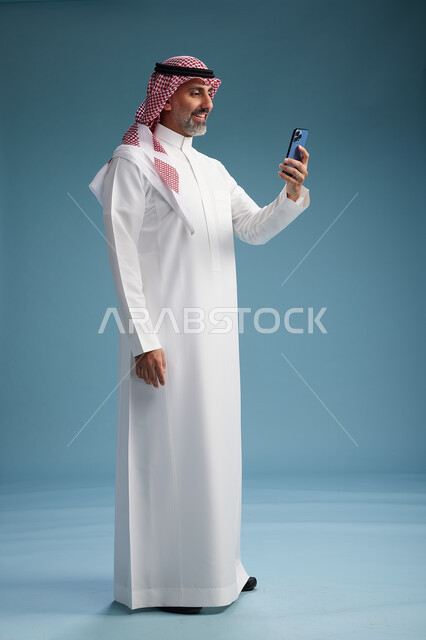 رجل  عربي سعودي خليجي يرتدي الثوب الابيض والشماغ  يستخدم هاتفاً ذكياً, الجسم بالطول الكامل, خلفيه زرقاء