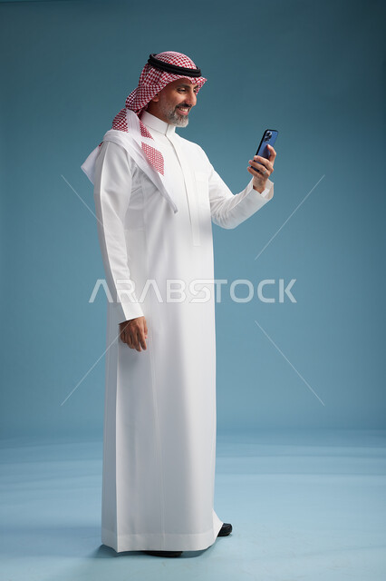 رجل  عربي سعودي خليجي يرتدي الثوب الابيض والشماغ  يستخدم هاتفاً ذكياً, الجسم بالطول الكامل, خلفيه زرقاء
