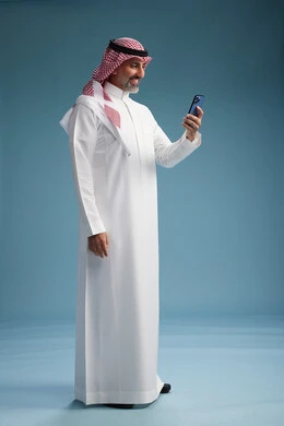 A Saudi Arabian Gulf Arab man wearing a white thobe and a shemagh is using a smartphone, full-body, with a blue background.