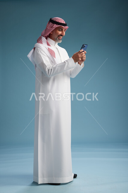 رجل  عربي سعودي خليجي يرتدي الثوب الابيض والشماغ  يستخدم هاتفاً ذكياً, الجسم بالطول الكامل, خلفيه زرقاء