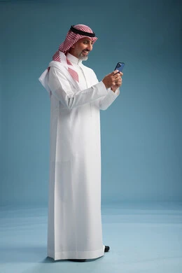 A Saudi Arabian Gulf Arab man wearing a white thobe and a shemagh is using a smartphone, full-body, with a blue background. A Saudi Arabian Gulf Arab man wearing a white thobe and a shemagh is using a smartphone, full-body, with a blue background.