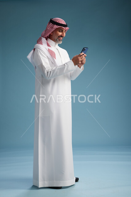 رجل  عربي سعودي خليجي يرتدي الثوب الابيض والشماغ  يستخدم هاتفاً ذكياً, الجسم بالطول الكامل, خلفيه زرقاء