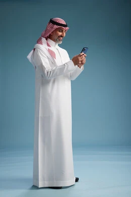 رجل  عربي سعودي خليجي يرتدي الثوب الابيض والشماغ  يستخدم هاتفاً ذكياً, الجسم بالطول الكامل, خلفيه زرقاء