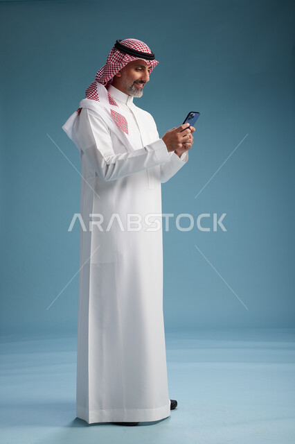 رجل  عربي سعودي خليجي يرتدي الثوب الابيض والشماغ  يستخدم هاتفاً ذكياً, الجسم بالطول الكامل, خلفيه زرقاء