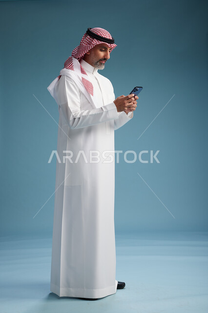رجل  عربي سعودي خليجي يرتدي الثوب الابيض والشماغ  يستخدم هاتفاً ذكياً, الجسم بالطول الكامل, خلفيه زرقاء