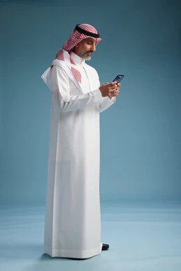 رجل  عربي سعودي خليجي يرتدي الثوب الابيض والشماغ  يستخدم هاتفاً ذكياً, الجسم بالطول الكامل, خلفيه زرقاء