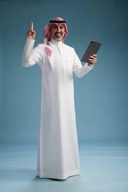بورتريه لرجل عربي سعودي  خليجي يرتدي الثوب  الابيض والشماغ يستخدم جهازاً لوحياً رقمياً, كامل الجسم, خلفيه زرقاء