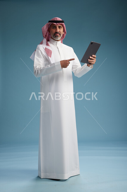 بورتريه لرجل عربي سعودي  خليجي يرتدي الثوب  الابيض والشماغ يستخدم جهازاً لوحياً رقمياً, كامل الجسم, خلفيه زرقاء