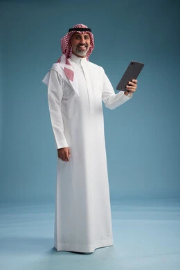 A portrait of a Saudi Arabian Gulf man wearing a white thobe and a shemagh using a digital tablet, full body, with a blue background.
