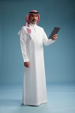 بورتريه لرجل عربي سعودي  خليجي يرتدي الثوب  الابيض والشماغ يستخدم جهازاً لوحياً رقمياً, كامل الجسم, خلفيه زرقاء