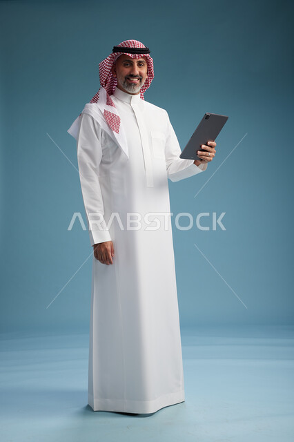 بورتريه لرجل عربي سعودي  خليجي يرتدي الثوب  الابيض والشماغ يستخدم جهازاً لوحياً رقمياً, كامل الجسم, خلفيه زرقاء