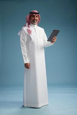 A portrait of a Saudi Arabian Gulf man wearing a white thobe and a shemagh using a digital tablet, full body, with a blue background.