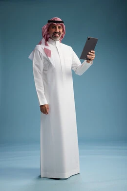 بورتريه لرجل عربي سعودي  خليجي يرتدي الثوب  الابيض والشماغ يستخدم جهازاً لوحياً رقمياً, كامل الجسم, خلفيه زرقاء