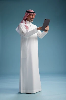A portrait of a Saudi Arabian Gulf man wearing a white thobe and a shemagh using a digital tablet, full body, with a blue background.