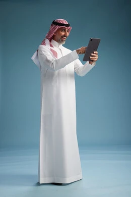 A portrait of a Saudi Arabian Gulf man wearing a white thobe and a shemagh using a digital tablet, full body, with a blue background.