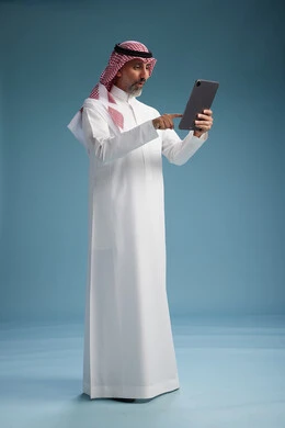 A portrait of a Saudi Arabian Gulf man wearing a white thobe and a shemagh using a digital tablet, full body, with a blue background.