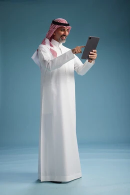 A portrait of a Saudi Arabian Gulf man wearing a white thobe and a shemagh using a digital tablet, full body, with a blue background.