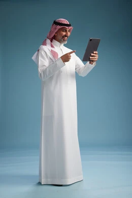 A portrait of a Saudi Arabian Gulf man wearing a white thobe and a shemagh using a digital tablet, full body, with a blue background.